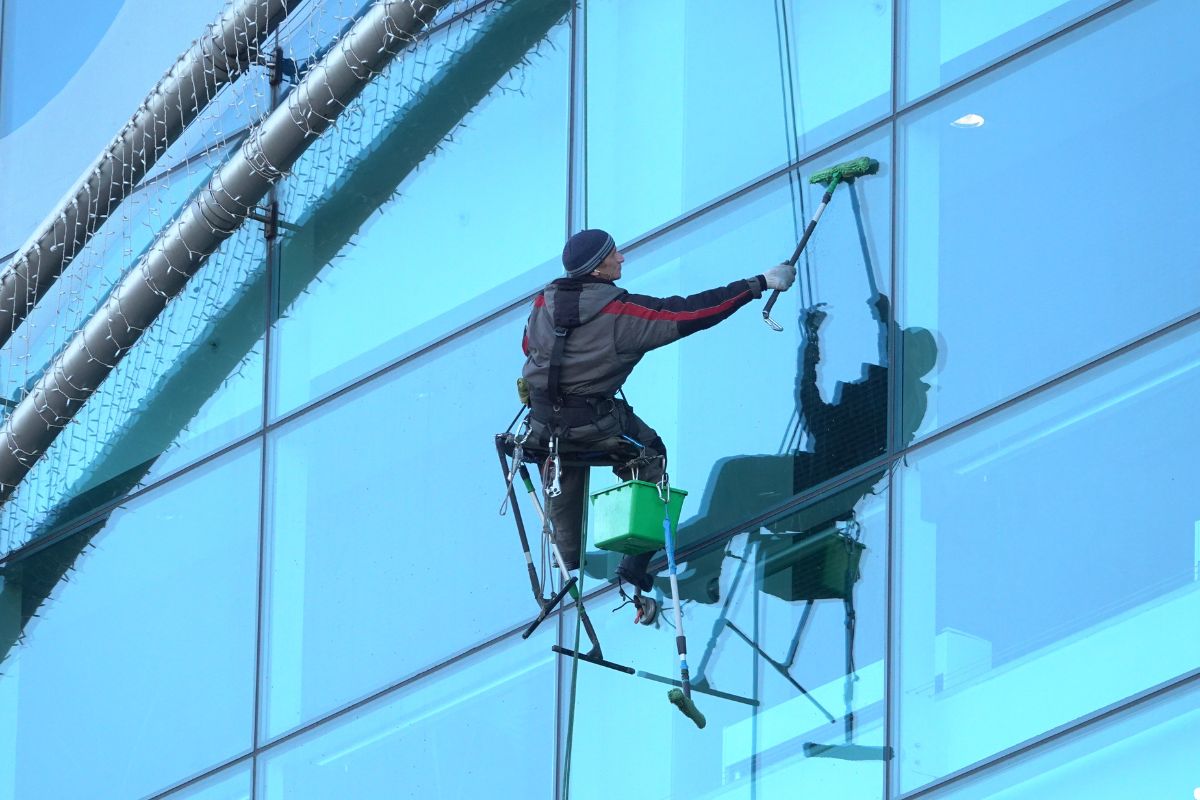A man in a safety harness cleans the glass exterior of a tall building with a squeegee and bucket.